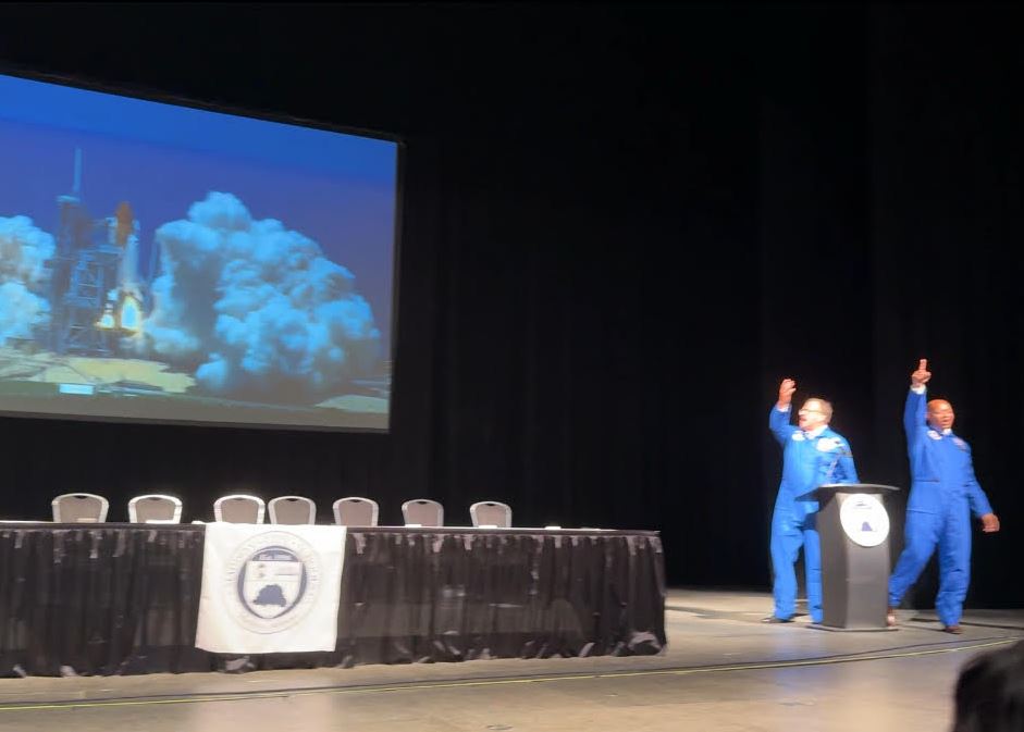  Dr. Nichols and Eric Terrell in space suits with rocket launching on screen behind them